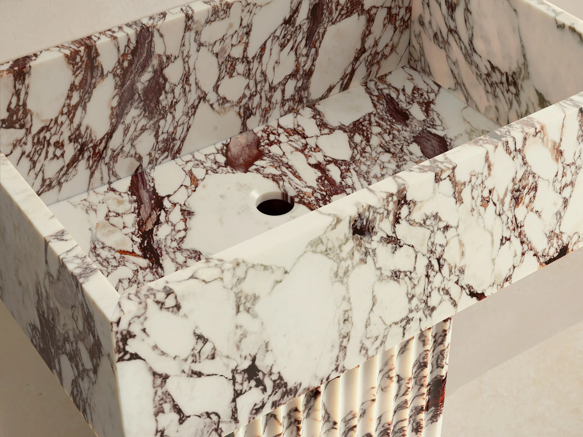 Front view of the Riven Pedestal Sink, a white marble sink with reddish-brown veining, a fluted pedestal base and rectangular basin.