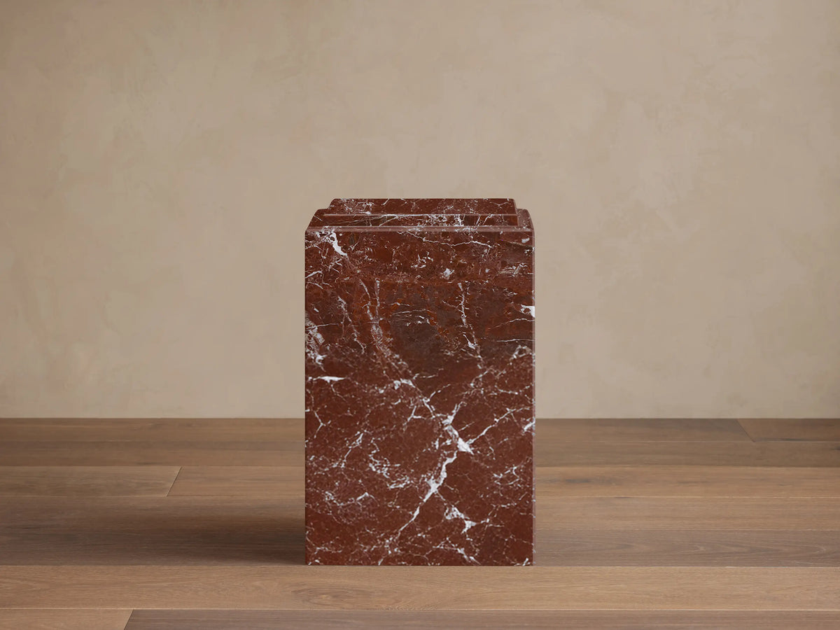 Front view of a reddish-brown Marmont Pedestal with natural white veining and a tiered top edge, standing on a wooden floor.