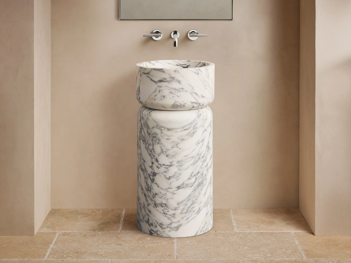 Front view of a white marble pedestal sink with prominent grey veining, featuring a rounded basin atop a cylindrical base.