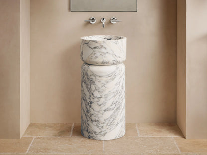 Front view of a white marble pedestal sink with prominent grey veining, featuring a rounded basin atop a cylindrical base.