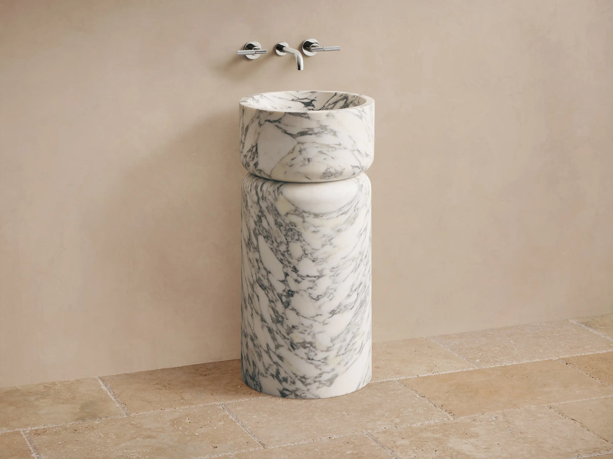 Front view of a white marble pedestal sink with prominent grey veining, featuring a rounded basin atop a cylindrical base.