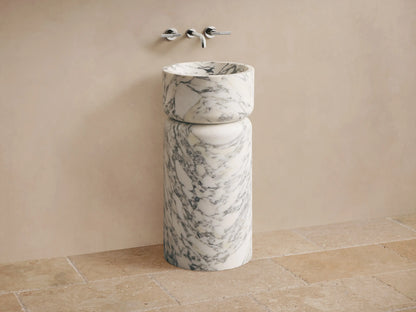 Front view of a white marble pedestal sink with prominent grey veining, featuring a rounded basin atop a cylindrical base.