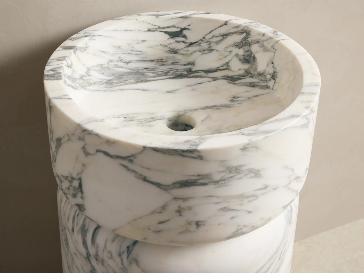 Front view of a white marble pedestal sink with grey veining, featuring a round basin on a cylindrical base. Wall-mounted faucets above.