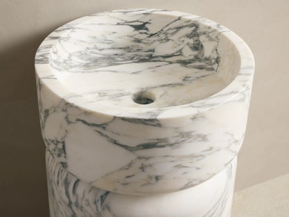 Front view of a white marble pedestal sink with grey veining, featuring a round basin on a cylindrical base. Wall-mounted faucets above.