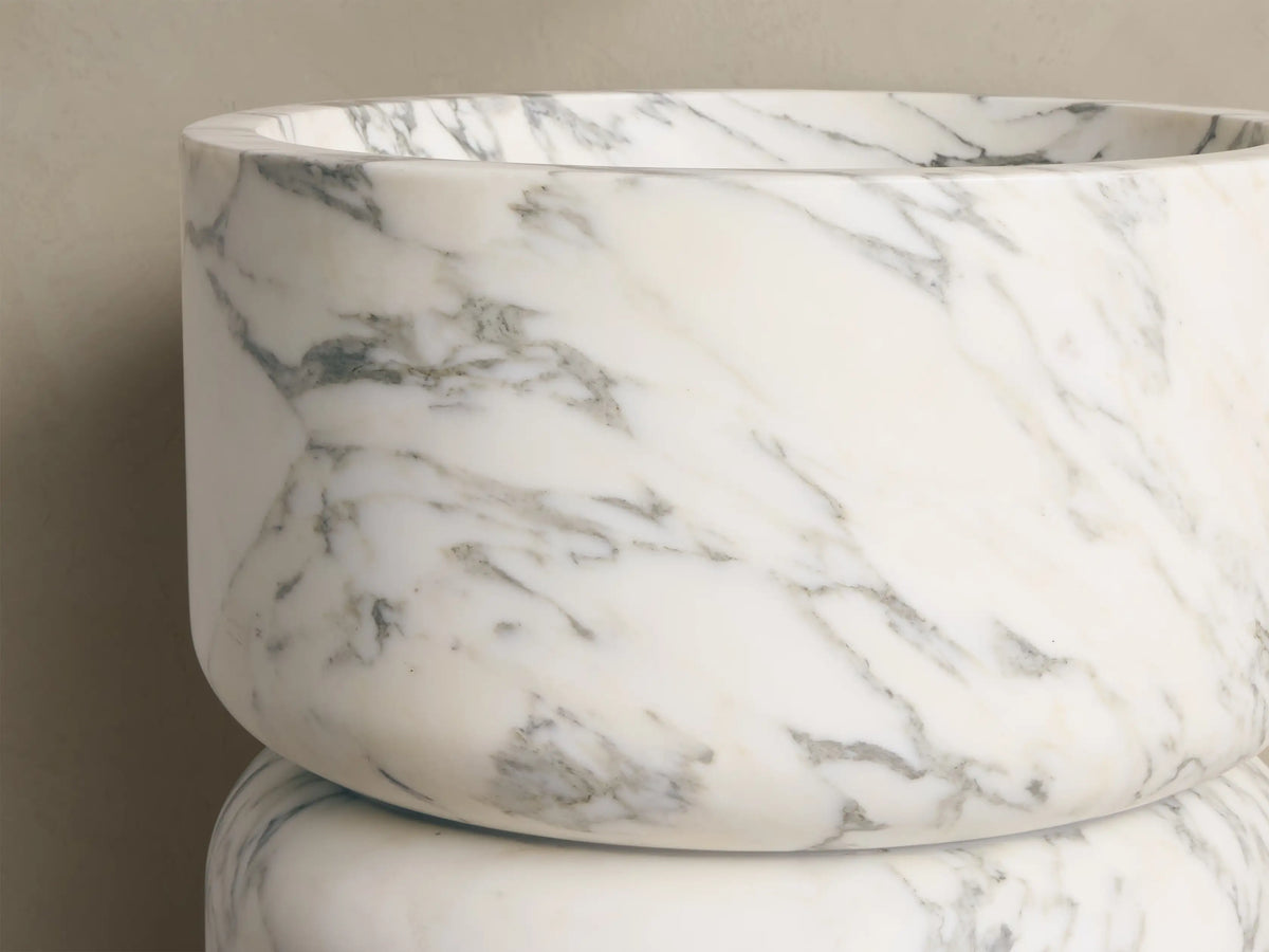 Angled view of a round white marble freestanding basin with dark gray veining, featuring an interior bowl and drain hole.