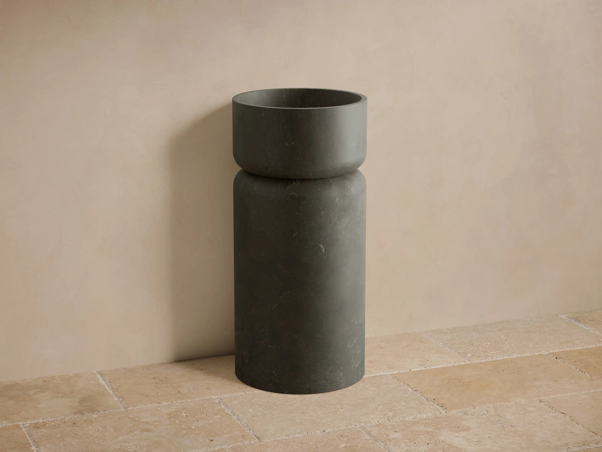 Front view of a dark gray Vaso Pedestal Sink, crafted from honed natural stone, with a cylindrical base and integrated bowl.