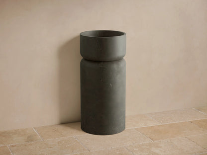 Front view of a dark gray Vaso Pedestal Sink, crafted from honed natural stone, with a cylindrical base and integrated bowl.