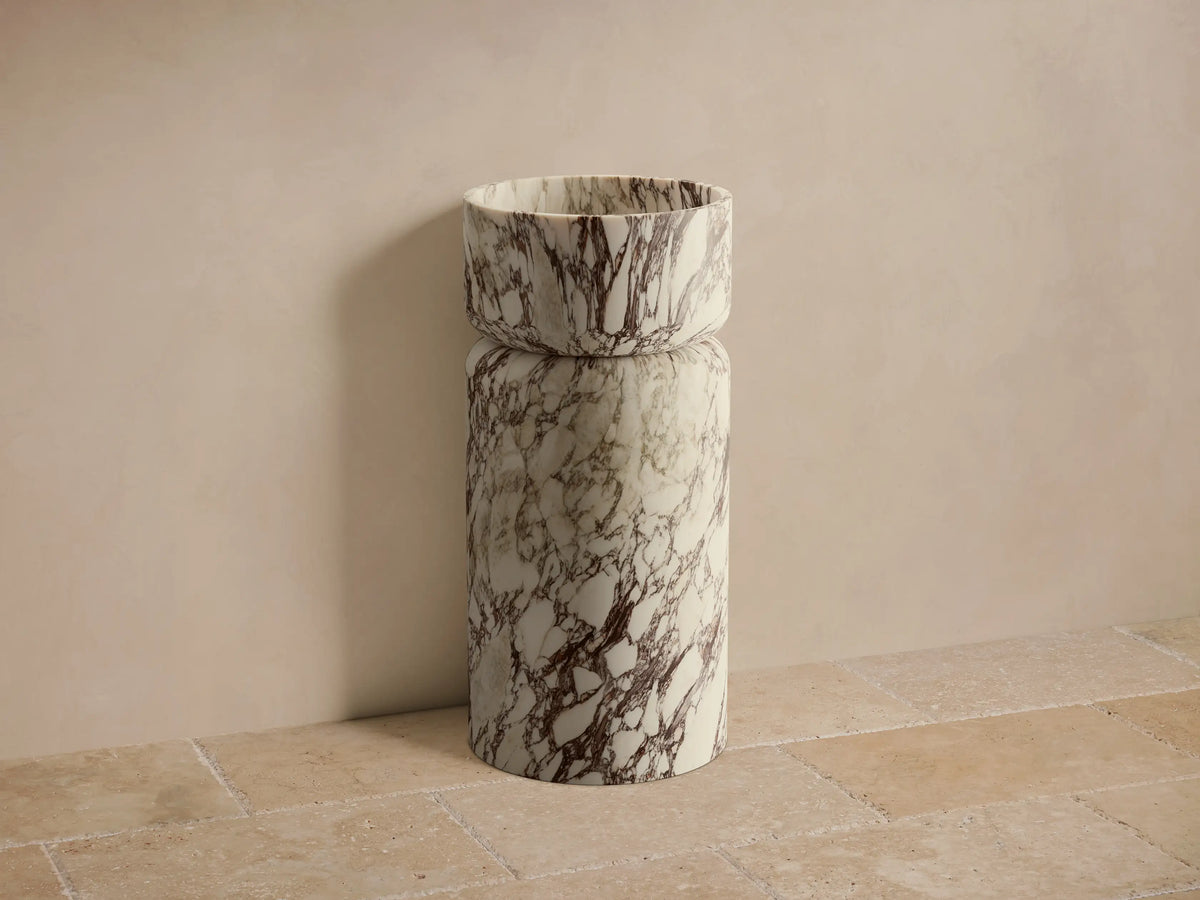 Front view of a white marble Vaso Pedestal Sink with dark brown veining, featuring a cylindrical base and integrated bowl.