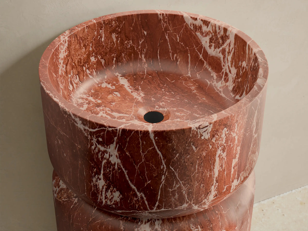 Front view of the Vaso Pedestal Sink, a reddish-brown marble pedestal with a cylindrical base and integrated basin, featuring white veining.