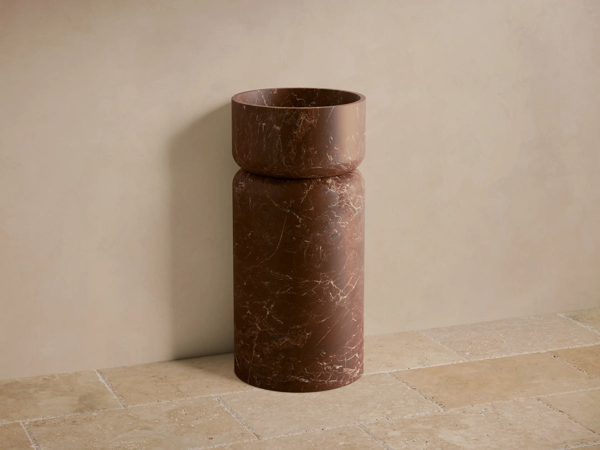 Front view of the dark reddish-brown Vaso Pedestal Sink, a cylindrical design with white veining on honed natural stone.