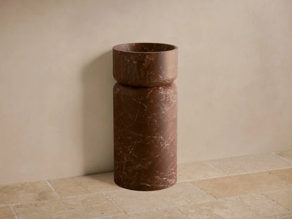 Front view of the dark reddish-brown Vaso Pedestal Sink, a cylindrical design with white veining on honed natural stone.