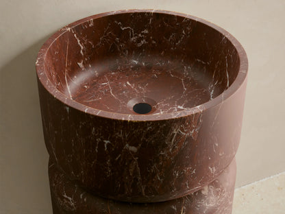 Front view of the reddish-brown marble Vaso Pedestal Sink, with white veining, featuring a cylindrical base and round basin.