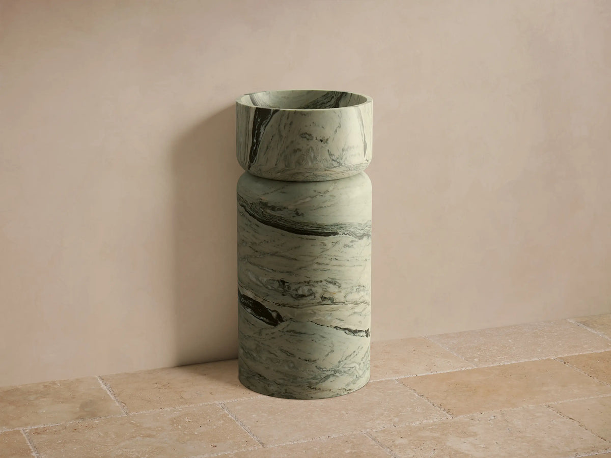 Front view of light green marble Vaso Pedestal Sink with dark gray and white veining, cylindrical base, and round integrated basin.