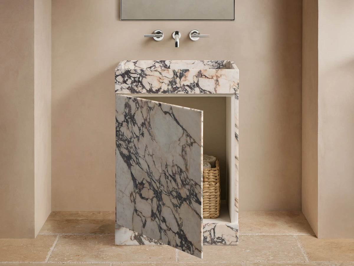 Front view of a white marble sink vanity with purple-black veining. Its door is ajar, revealing a woven basket with towels.