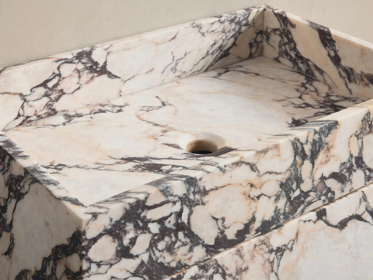 Front view of white Marble Pedestal Sink with black and pink veining and square basin. Chrome wall-mounted faucet.