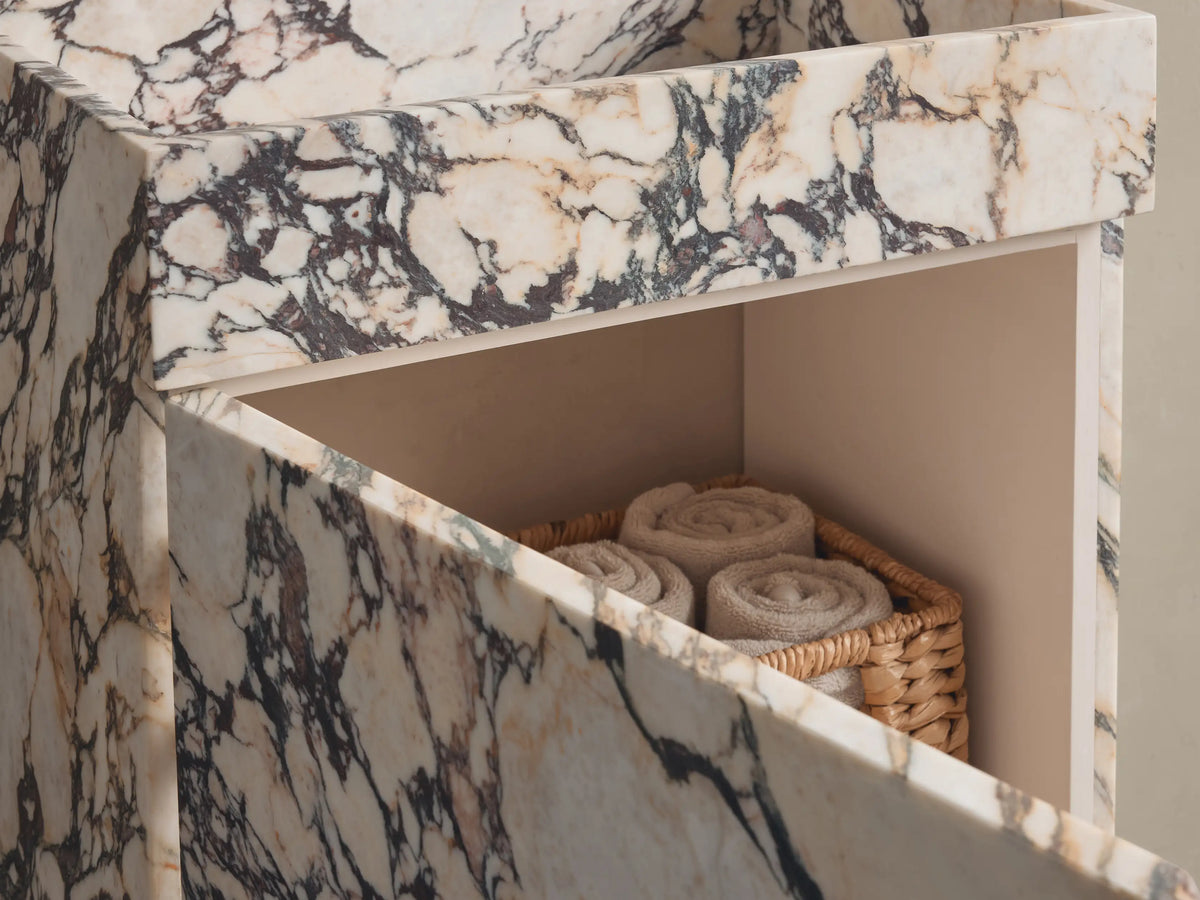 Close-up of a rectangular marble basin. Cream and light brown marble with prominent dark gray and black veining, showing a drain.
