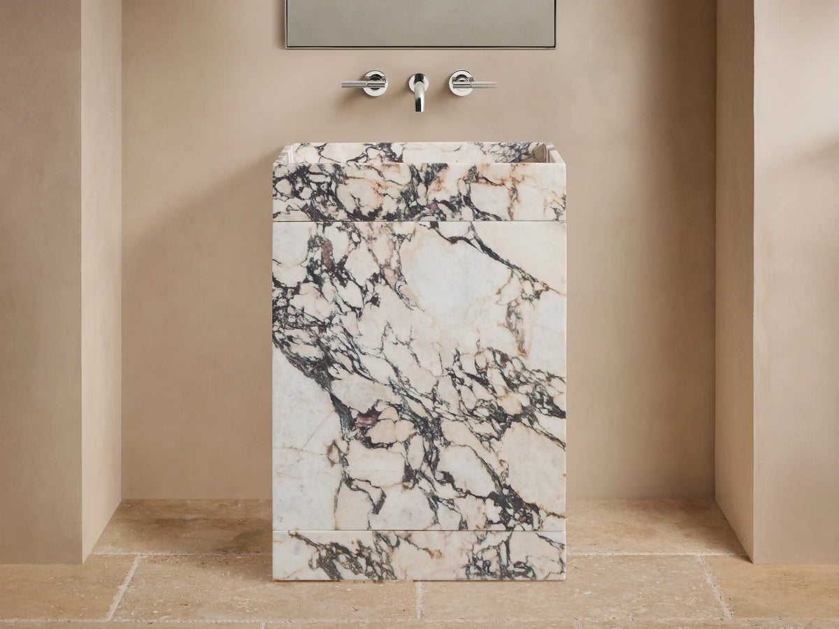 Open drawer view of a cream marble vanity with dark brown veining, revealing a woven basket holding rolled cream towels.