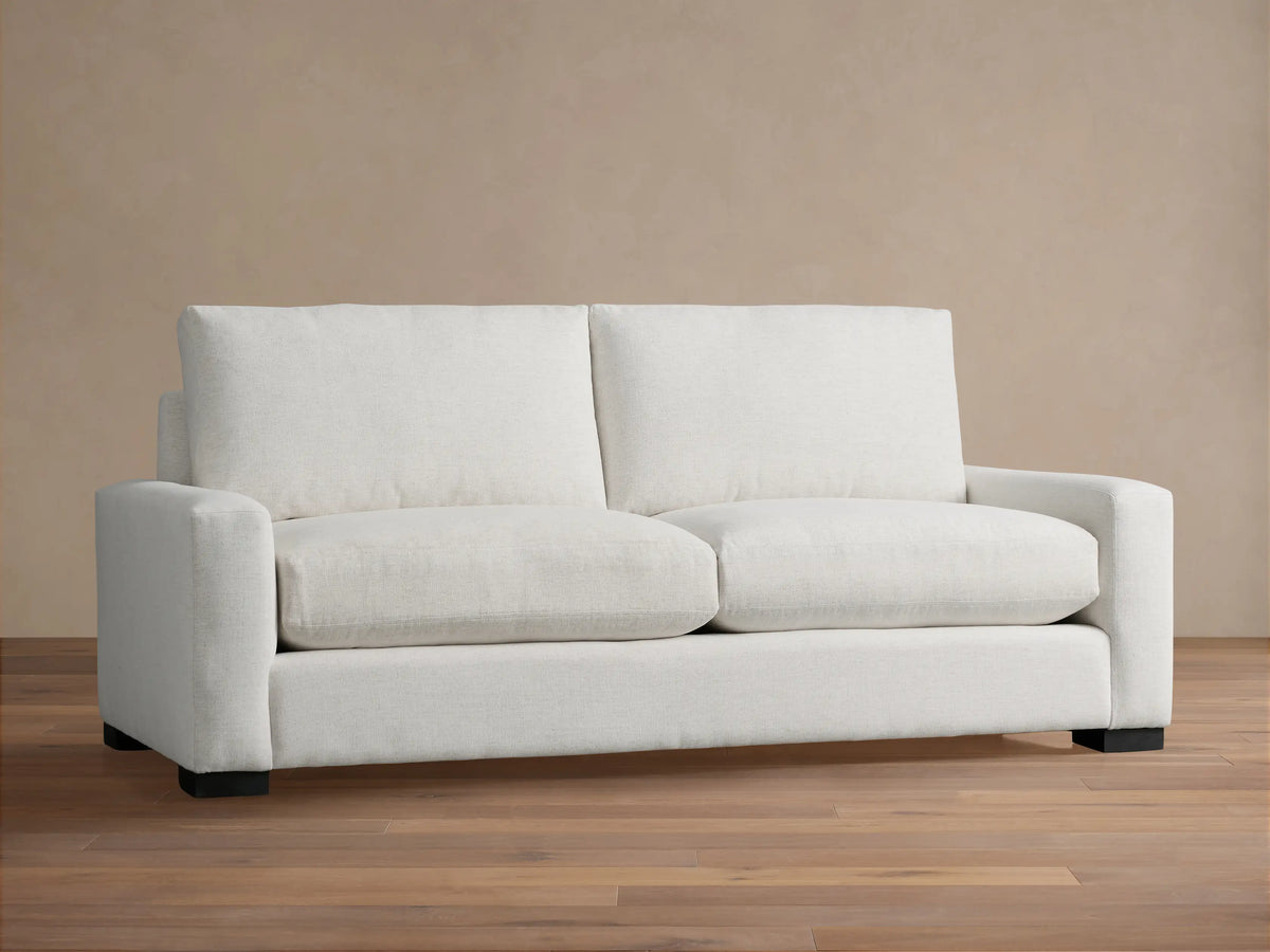 Remington Sofa, a modern off-white two-seater sofa with square arms and plush cushions, set against a warm tan wall.