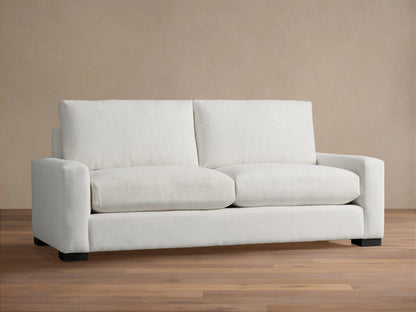 Remington Sofa, a modern off-white two-seater sofa with square arms and plush cushions, set against a warm tan wall.