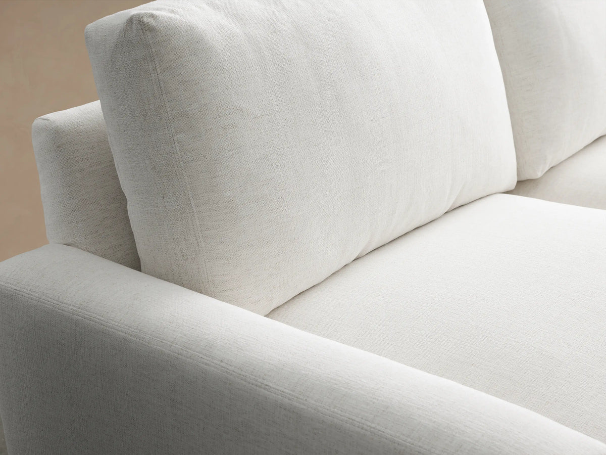 Remington Sofa, 2-seater, light grey fabric, modern design, thick cushions, square arms, on a wooden floor against a beige wall.