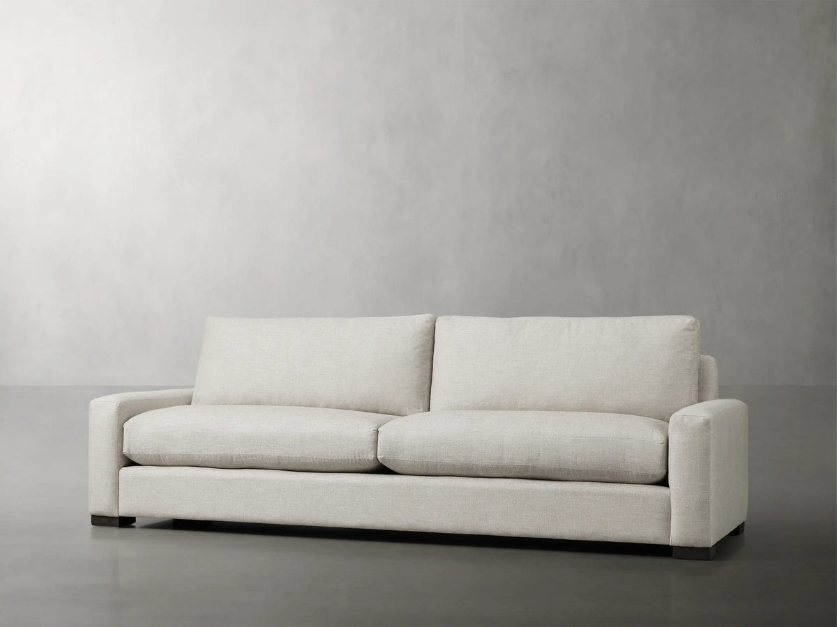 Front view of Remington Sofa, upholstered in light beige fabric, with two seat & back cushions, wide track arms, and dark wood feet.