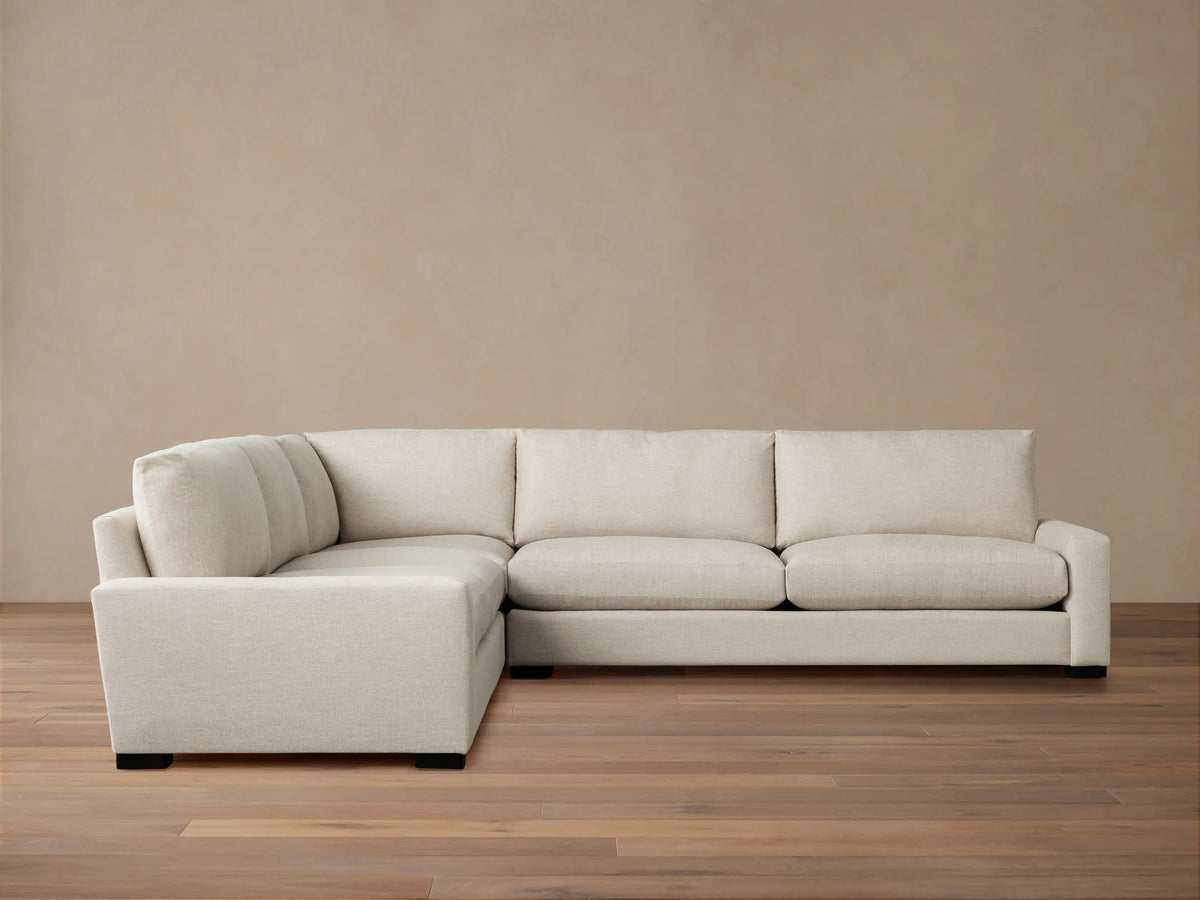 Off-white Remington Three-Piece Corner Sectional, an L-shaped sofa with plush cushions, against a warm beige wall & wood floor.