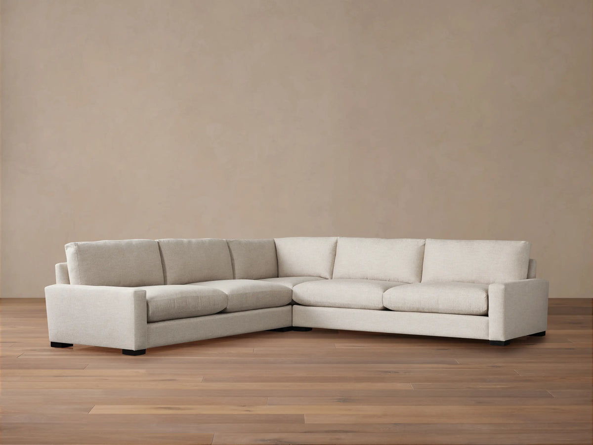 Off-white Remington Three-Piece Corner Sectional, an L-shaped sofa with plush cushions, against a warm beige wall & wood floor.