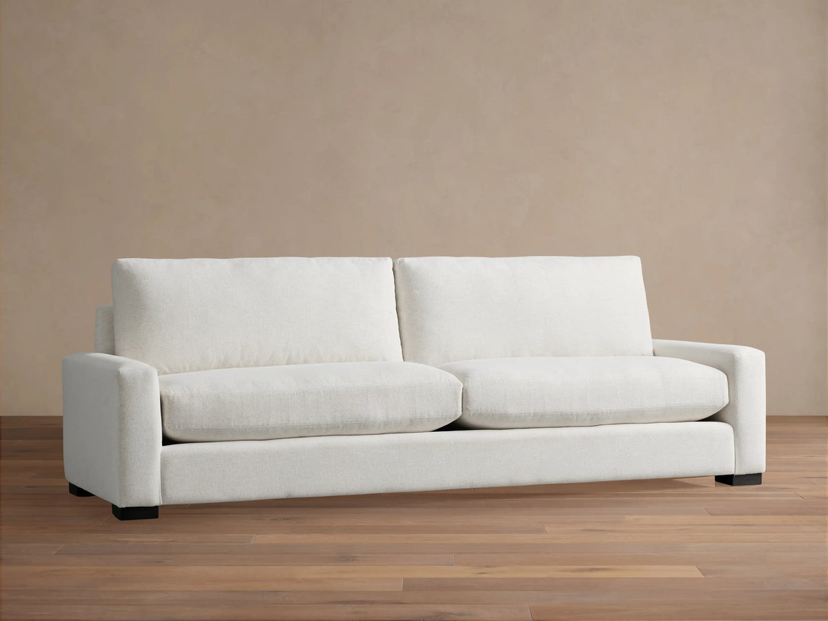 Remington Sofa, a modern off-white 2-seater sofa with plush cushions and wide arms, on a wooden floor against a beige wall.