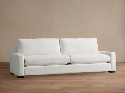 Remington Sofa, a modern off-white 2-seater sofa with plush cushions and wide arms, on a wooden floor against a beige wall.