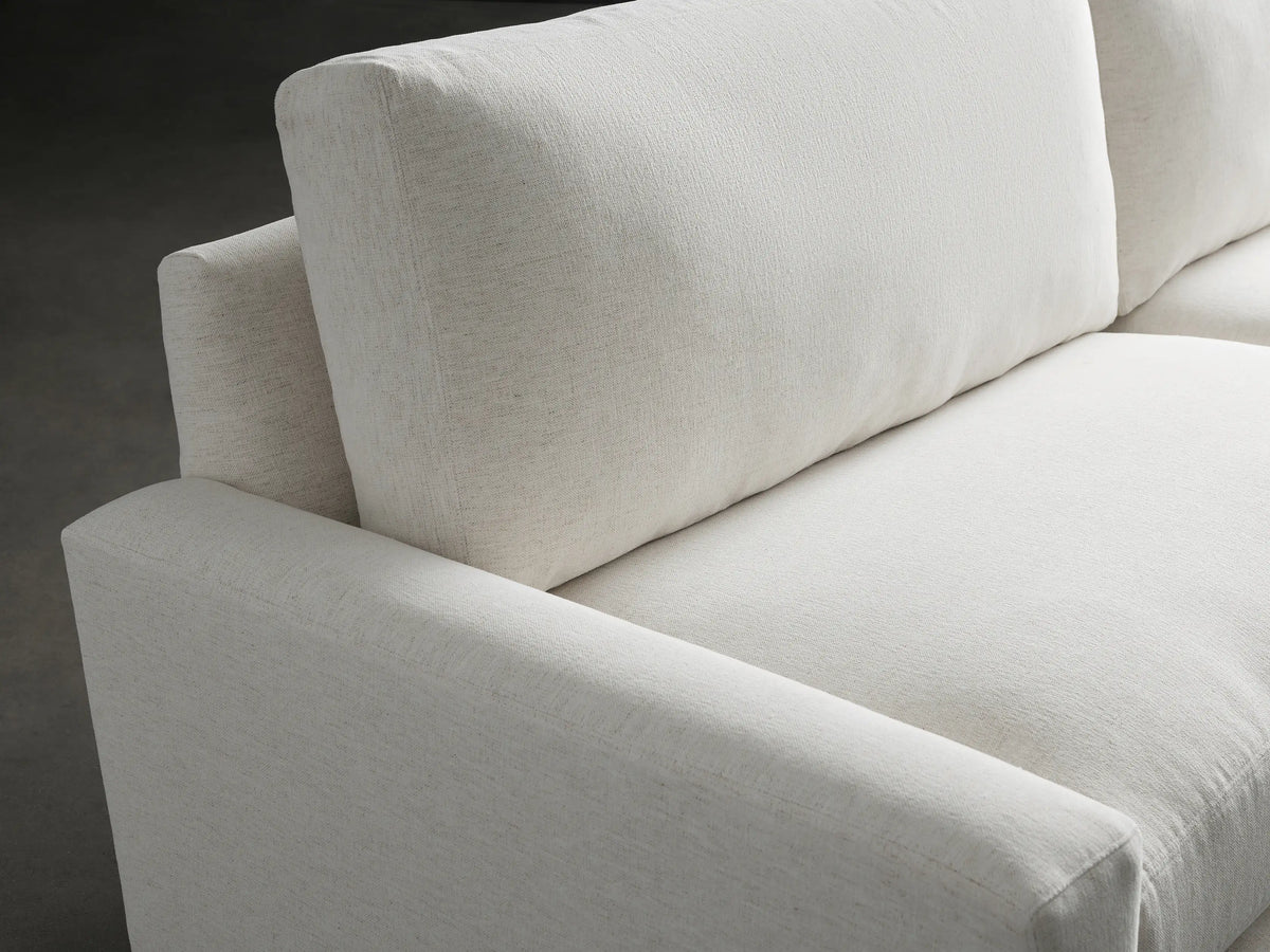 Remington Sofa: a cream-colored, low-profile two-seater sofa with wide arms and textured fabric, angled slightly on a gray floor.