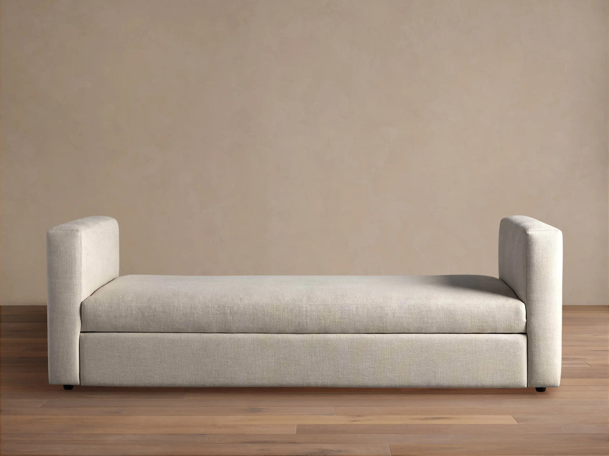 Light beige Remington Trundle Sleeper Daybed with two low, rounded armrests, on a wooden floor against a beige wall.