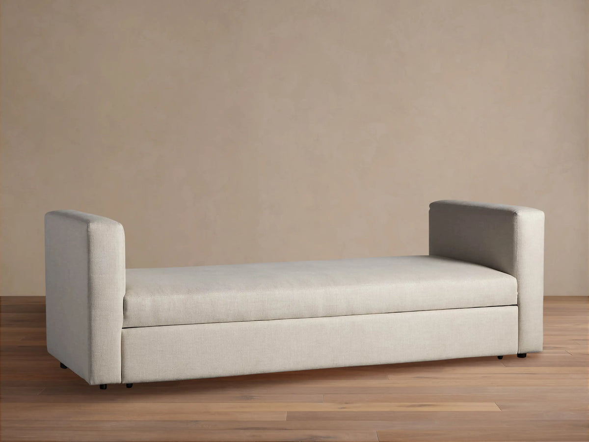 Light beige Remington Trundle Sleeper Daybed with two low, rounded armrests, on a wooden floor against a beige wall.
