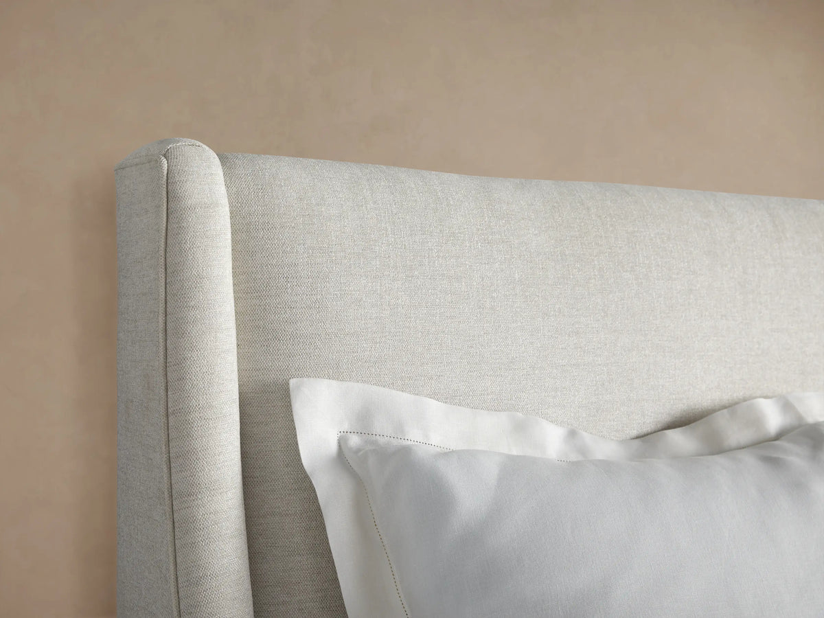 Front view of a light beige upholstered Atlyn Bed, featuring a tall, slightly winged headboard, and dressed with white pillows and duvet.