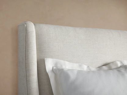Front view of a light beige upholstered Atlyn Bed, featuring a tall, slightly winged headboard, and dressed with white pillows and duvet.