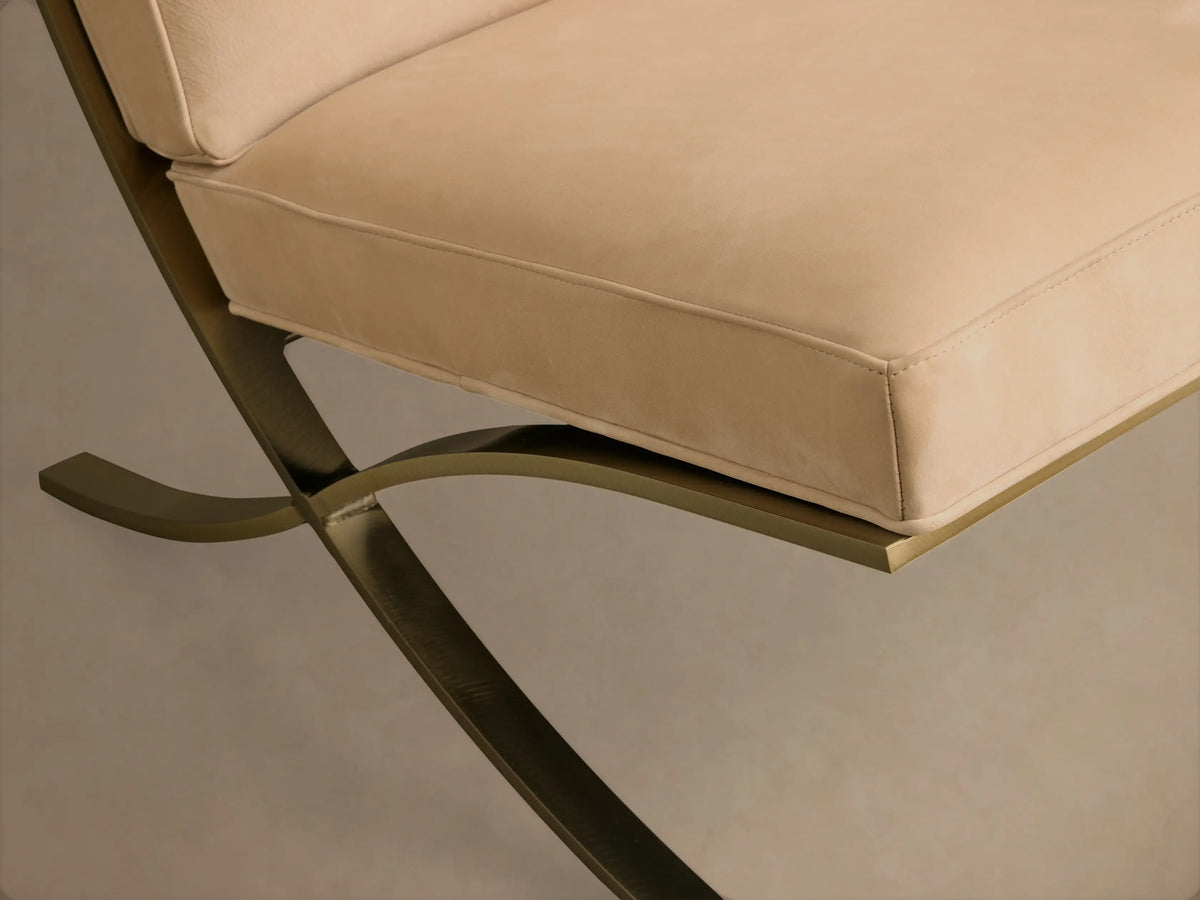 Close-up of the light beige leather seat cushion with piped edges and the bronze metallic X-shaped base of the Moxie Leather Chair.
