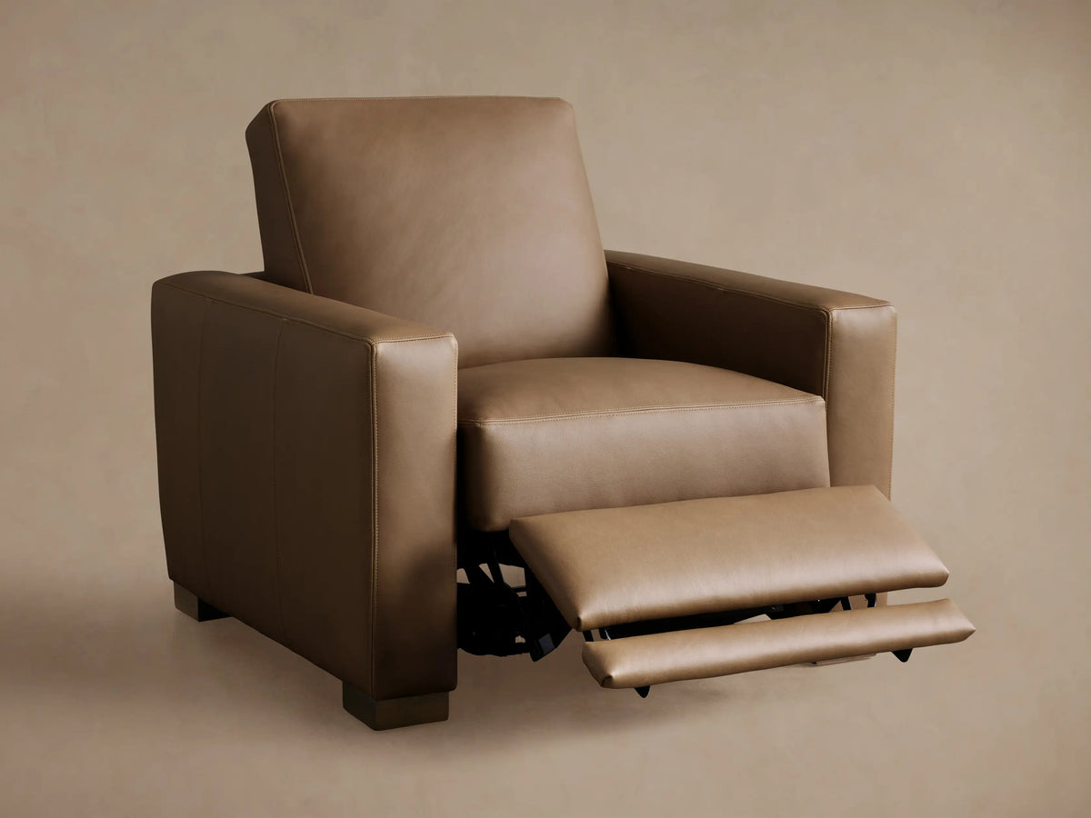 3/4 rear view of a tan leather Remington Motion Recliner with clean stitching, power port, and dark wood block feet.