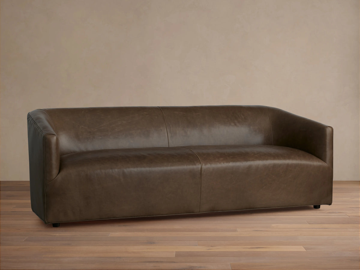 Brown Ellison Leather Sofa with a sleek, modern design. Features smooth, continuous curves and a slight sheen, set on a wooden floor.