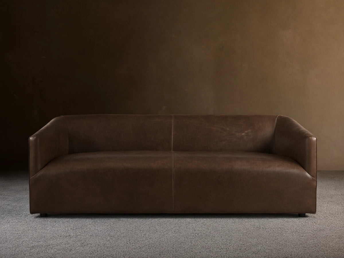 Brown Ellison Leather Sofa with a sleek, modern design. Features smooth, continuous curves and a slight sheen, set on a wooden floor.