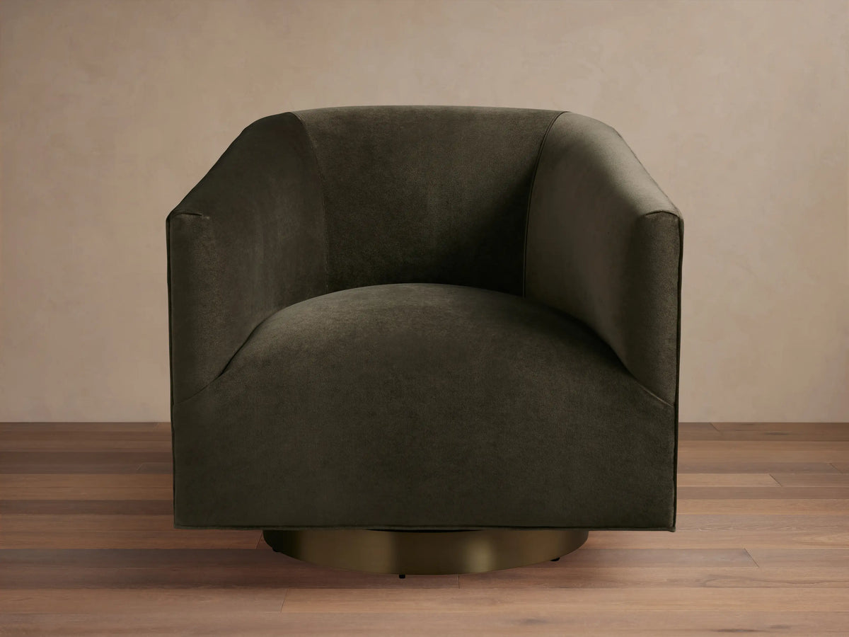 Dark brown Ellison Metal Plinth Swivel Chair with a rounded, cushioned backrest and armrests, on a brass-toned swivel base, against a wooden floor.