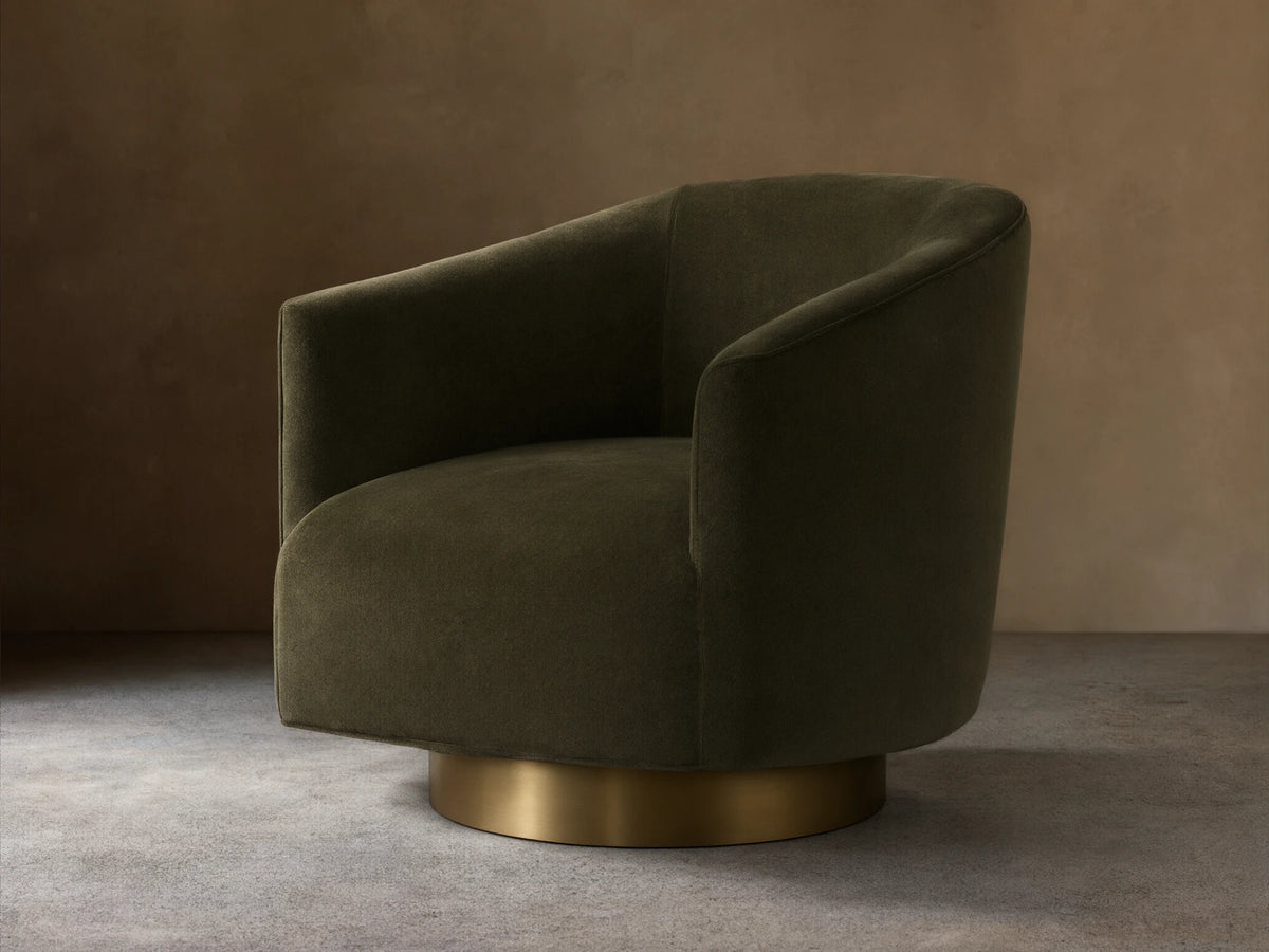 Dark brown Ellison Metal Plinth Swivel Chair with a rounded, cushioned backrest and armrests, on a brass-toned swivel base, against a wooden floor.