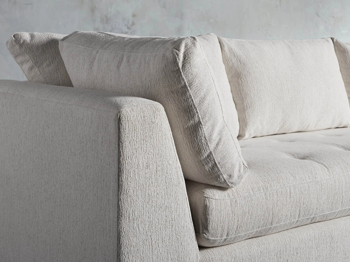 Light beige Bryden Two-Piece Daybed Sectional, L-shaped with dark wood base, against a textured grey wall.