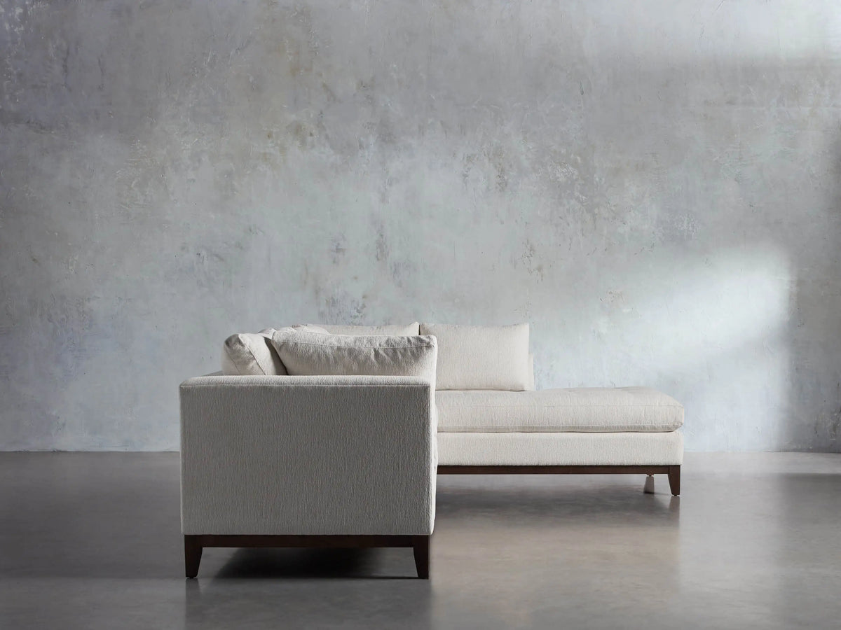 Close-up of the Bryden Two-Piece Daybed Sectional's light textured fabric, dark wooden base, and tapered leg on a gray floor.