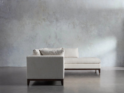 Close-up of the Bryden Two-Piece Daybed Sectional's light textured fabric, dark wooden base, and tapered leg on a gray floor.