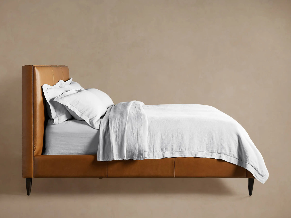 Close-up of Andes Leather Bed. Brown leather frame with dark tapered wooden leg. Light grey blanket draped over.
