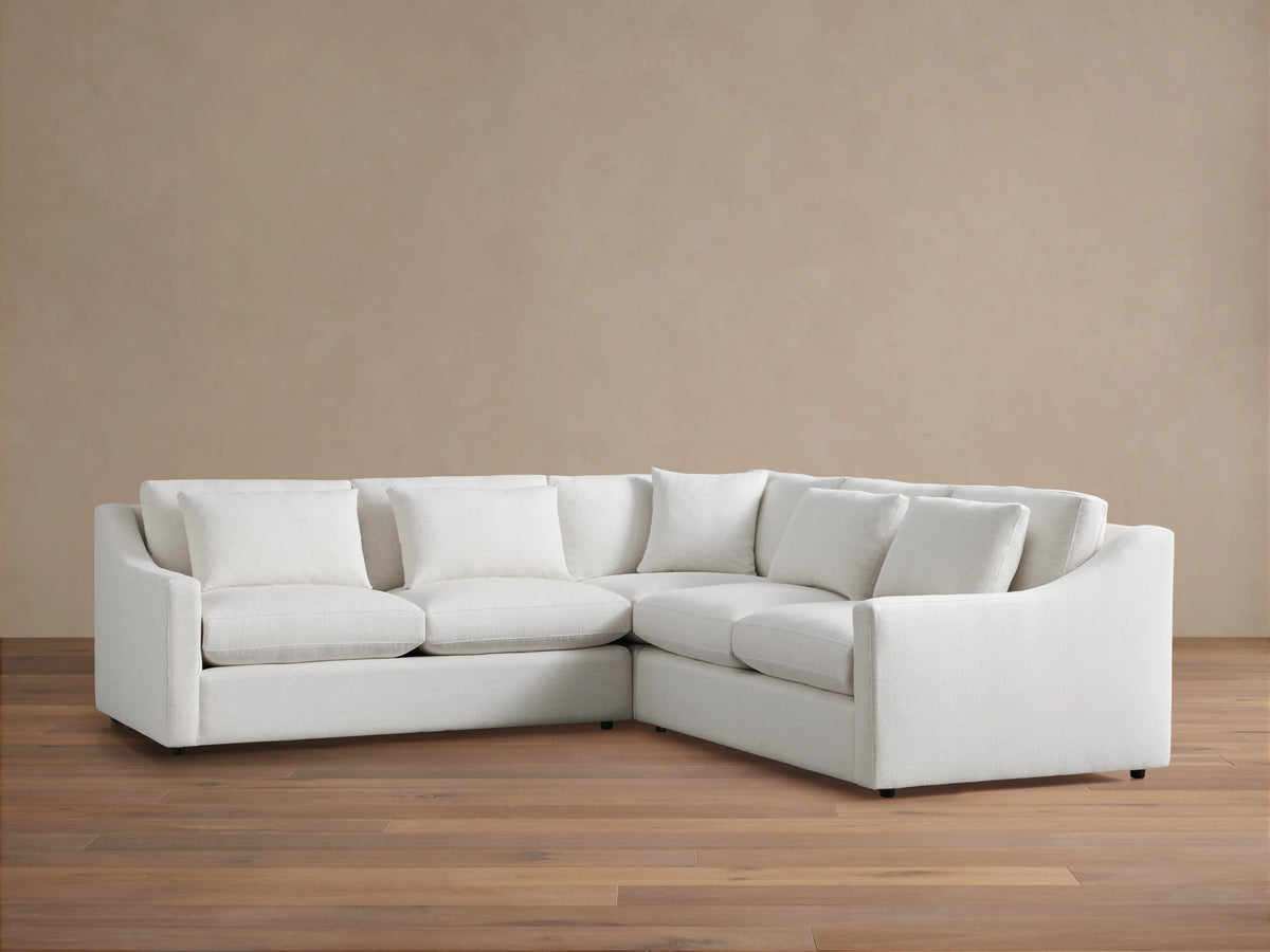 Alt text: Ashby Three-Piece Corner Sectional in off-white fabric, featuring three seat cushions and backrests, displayed on a wooden floor against a beige wall.
