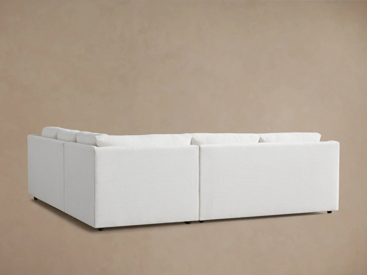 Corner of the Ashby Three-Piece Sectional in a soft, light fabric, featuring plump back cushions and rounded armrest.