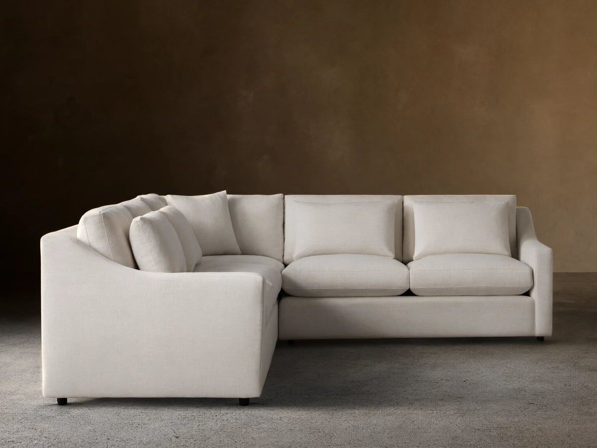 Alt text: Ashby Three-Piece Corner Sectional in off-white fabric, featuring three seat cushions and backrests, displayed on a wooden floor against a beige wall.