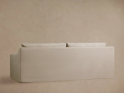 Ashby Slipcovered Sofa in a neutral light beige woven fabric, featuring plush seat and back cushions, with a gently sloped armrest.