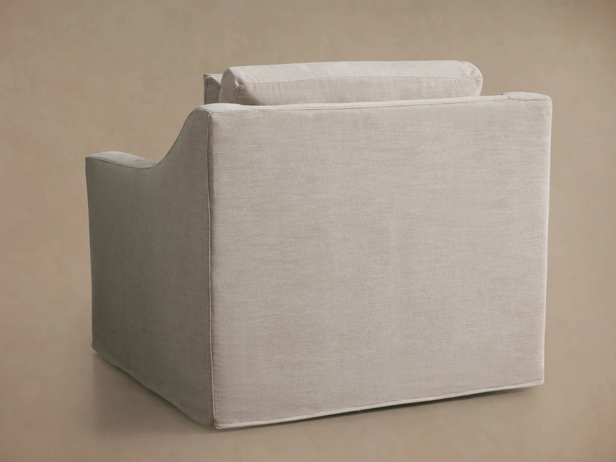 Ashby Slipcovered Swivel Chair with a light beige fabric, shown from the back at an angle. The chair has a boxy shape and visible cushion seams.