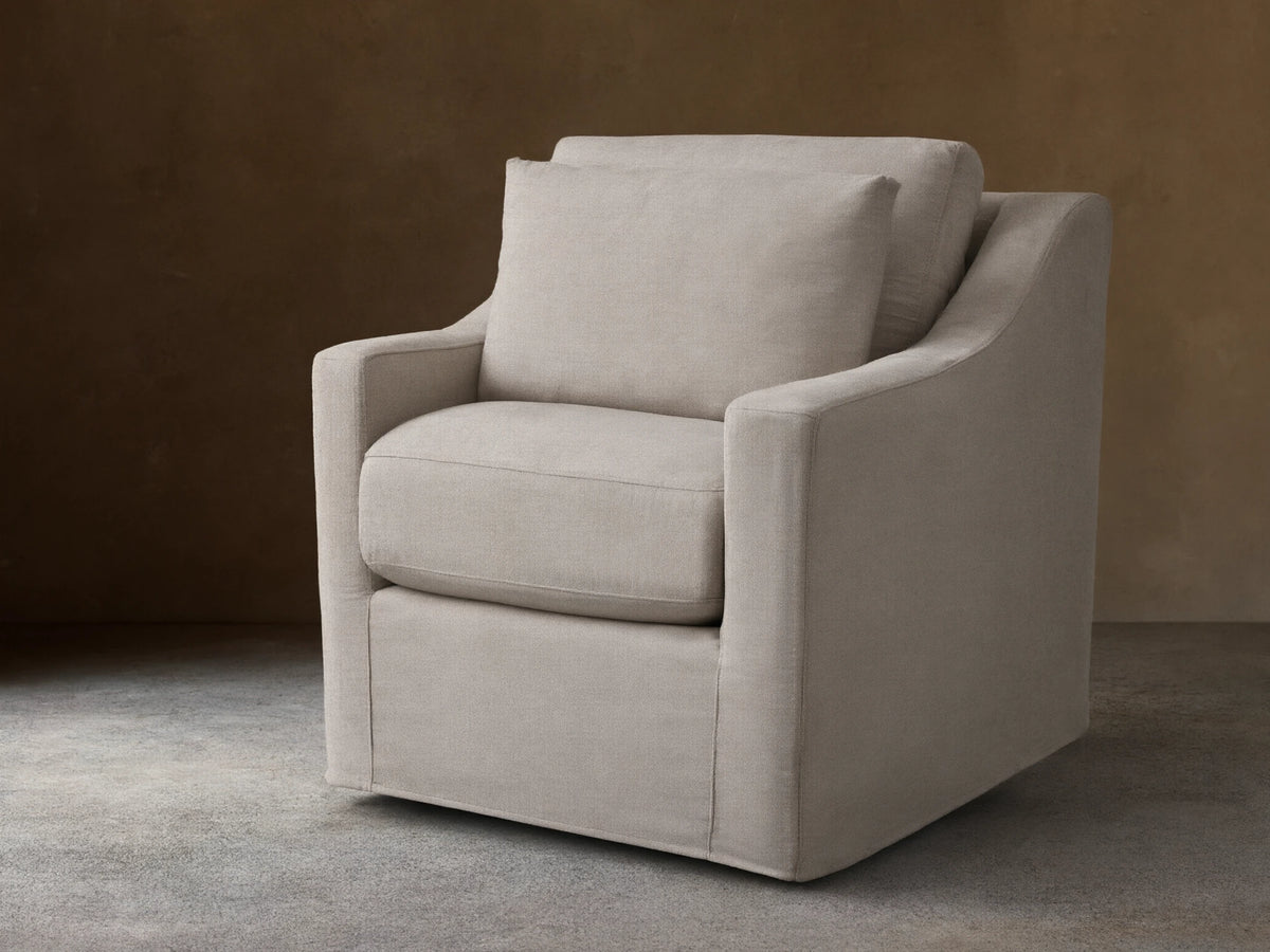 Ashby Slipcovered Swivel Chair in beige fabric on a wooden floor, featuring plush seat and back cushions, with a modern, rounded arm design.